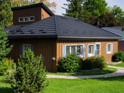 Wood siding multi-level home can be seen with a Black Hy-Grade Steel roof on a sunny day with blue sky. There are lots of plants and bushes in various colours of green surround the home.