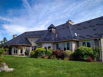 Hy-grade steel roof on a beige brick bungalo with green grass in the foreground and big bushes accent the garden in front of the home.