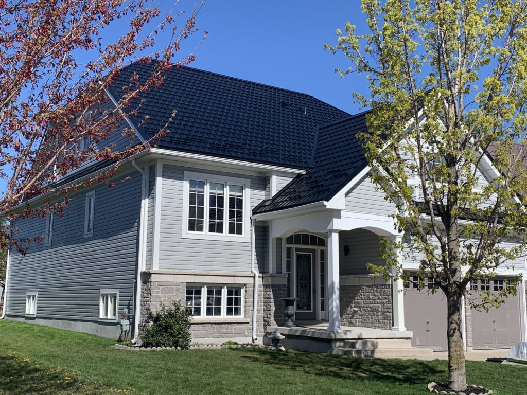 Hy-Grade Steel and Metal Roof in the colour Black. The 2-story house has grey siding with a sunny blue-sky background. There is bright green grass in front of the home.