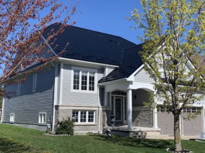Hy-Grade Steel and Metal Roof in the colour Black. The 2-story house has grey siding with a sunny blue-sky background. There is bright green grass in front of the home.