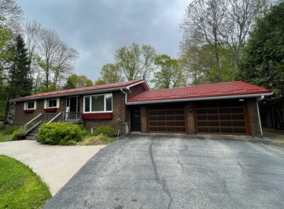 Hy-Grade-Steel-Roofing-System-Metal-Roofing-See-Our-Work-Tile-Red-bungalow-style house-with green grass and blue sky with dark brown brick sizing and brown garage doors. A long driveway is empty on the right.