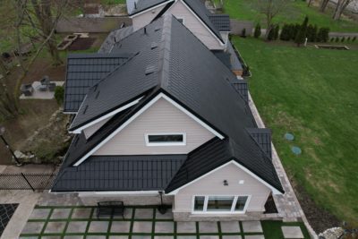 Hy-Grade Steel roof from above on a house with grey siding and white accents. There are lots of white patio stones around the home. A farmer's field can be seen off to the left in the background.