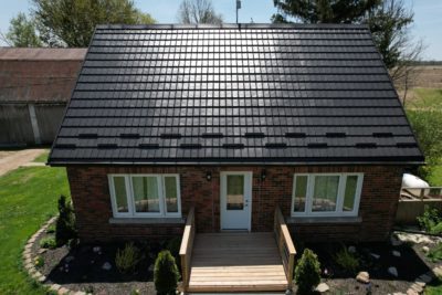 Bungalow home with a Black Hy-Grade Steel roof in a sunny day with blue sky. the angle is bird's eye from a drone and the sun is reflecting off the front of the home. The home has red brick.