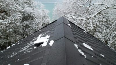 A Hy-Grade Steel Roof is seen with snow on top. Water can be see dripping off the roof and snow covered trees are in the background along with a blue lake view