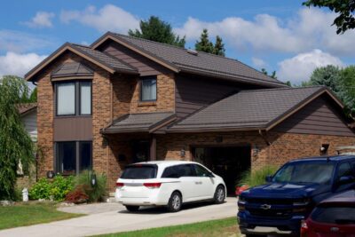 Hy-Grade Metal Roof on brown brick house with asphalt driveway and green garden to the left. A white minivan is in the driveway.