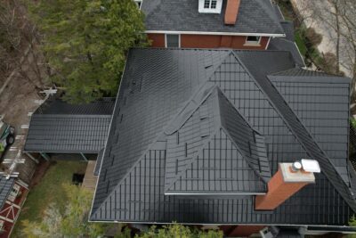 A Hy-Grade steel roof from a bird's eye view can be seen with a red brick chimney. Around the home is green trees and grass. Another home with an asphalt roof can be seen with similar red bricks in the background