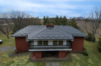 2 story red brick House with a walk-out balcony with black railing can be seen with a Black Hy-Grade Steel roof on a sunny day with blue sky.