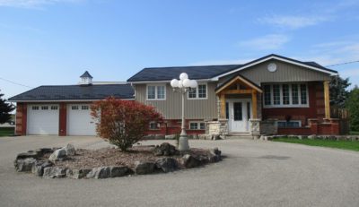 Hy-Grade Steel and Metal Roof in the colour Black. The bungalow house has grey siding with a sunny blue-sky background. There is a large asphalt driveway with a garden in the middle.