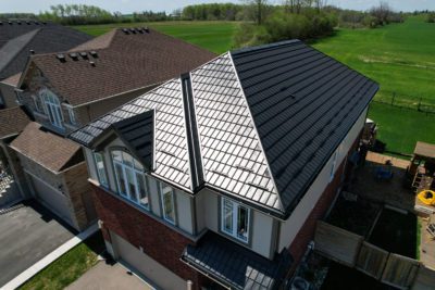 2 story house with a Black Hy-Grade Steel roof in a sunny day with blue sky. the angle is bird's eye from a drone and the sun is reflecting off the front of the home. The home has both red brick and white siding.