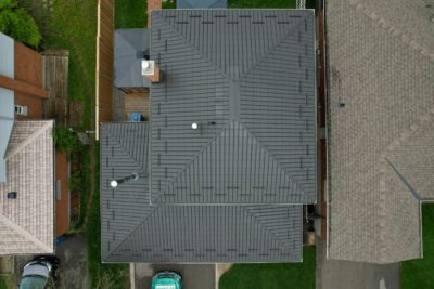 Drone angle bird's eye with Hy-Grade metal roof in dark brown. The top of a hy-grade steel truck is on the left of the driveway. A chimney is seen in the top left of the roof.