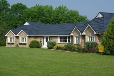 Beige siding bungalow home can be seen with a Black Hy-Grade Steel roof on a sunny day with blue sky. There are lots of plants and bushes in various colours of green surround the home.