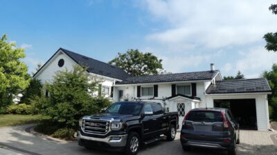 Hy-grade steel roof in black with two cars in the driveway, one a truck and one an SUV. The house is white with black accents. a sunny blue sky with clouds can be seen in the behind the home.